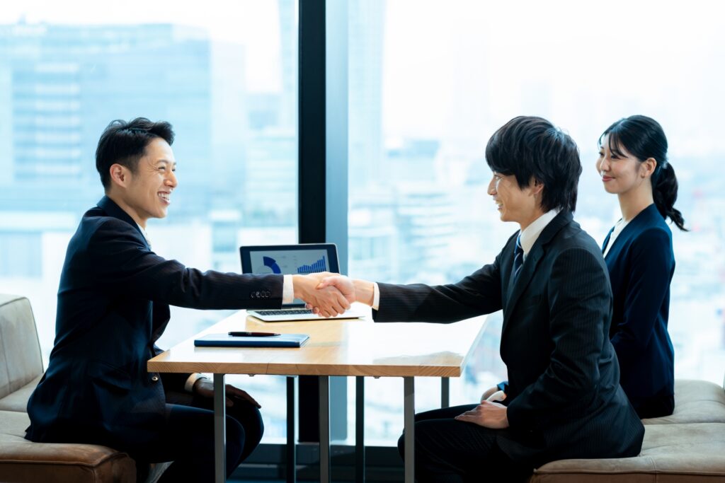 オフィスで握手を交わすビジネスパーソンと、データを表示したノートパソコン。業務支援SaaSの商談・提案シーン