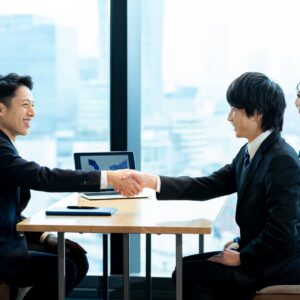 オフィスで握手を交わすビジネスパーソンと、データを表示したノートパソコン。業務支援SaaSの商談・提案シーン