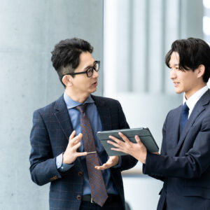 オフィスでタブレットを手に先輩社員と話す若手の営業担当者