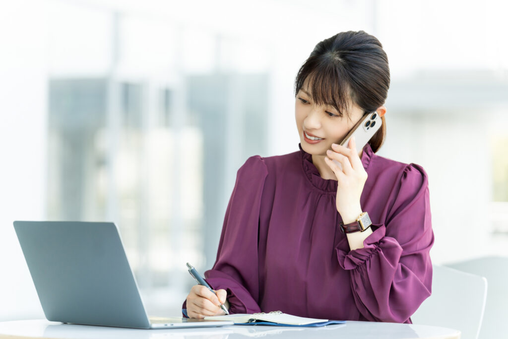 スマートフォンで通話しながらノートにメモを取る女性の営業担当者