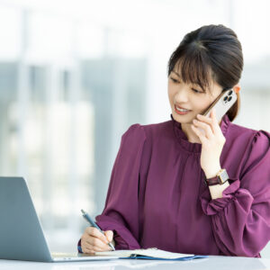 スマートフォンで通話しながらノートにメモを取る女性の営業担当者